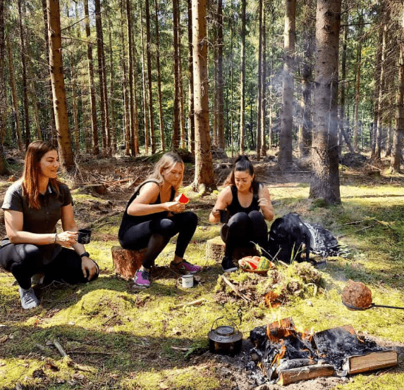 tre kvinder sidder i skovbunden og griller mad over baal
